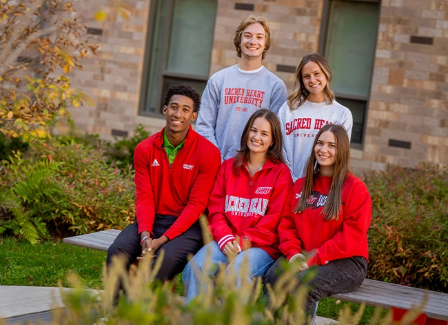 Students wearing Sacred Heart University apparel sitting and standing outdoors on campus
