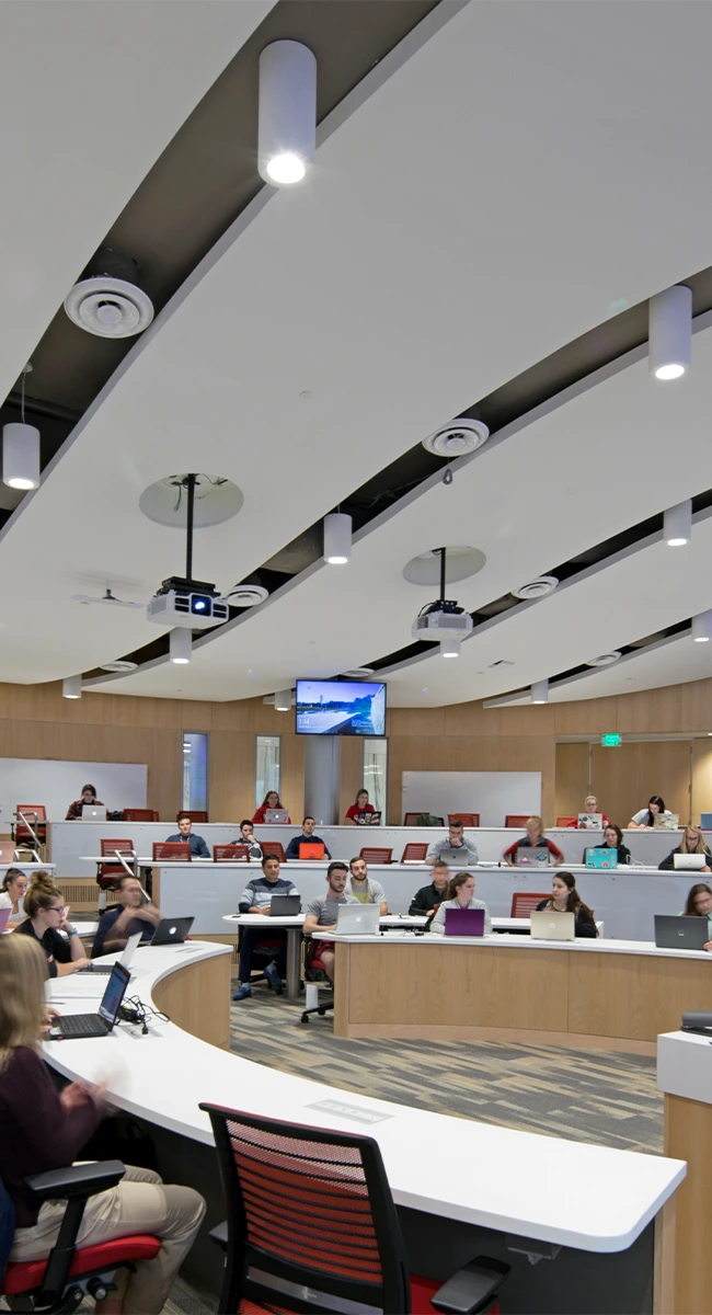 Students seated in a tiered lecture hall classroom using laptops.