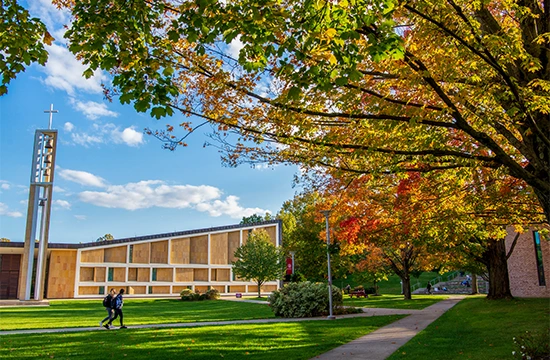 Church building with cross tower beside a lawn and walkway, surrounded by autumn trees