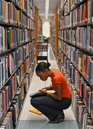 Student reading a book in a library aisle between shelves