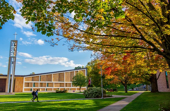 Church building with cross tower beside a lawn and walkway, surrounded by autumn trees